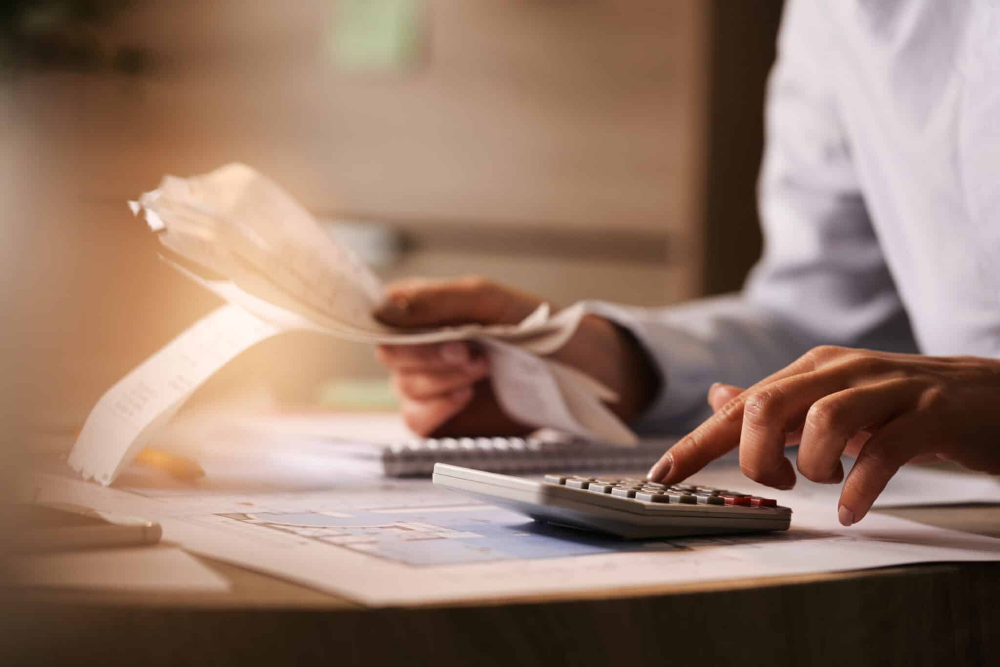 closeup economist using calculator while going through bills taxes office scaled