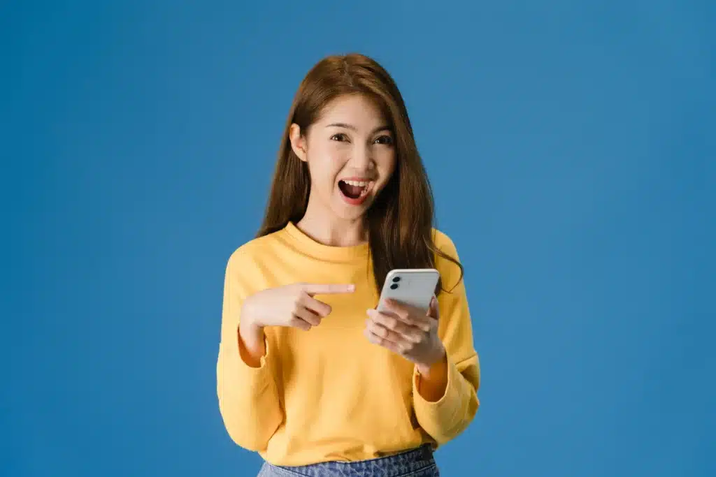 surprised young asia lady using mobile phone with positive expression smiles broadly dressed casual clothing looking camera blue background happy adorable glad woman rejoices success Web H